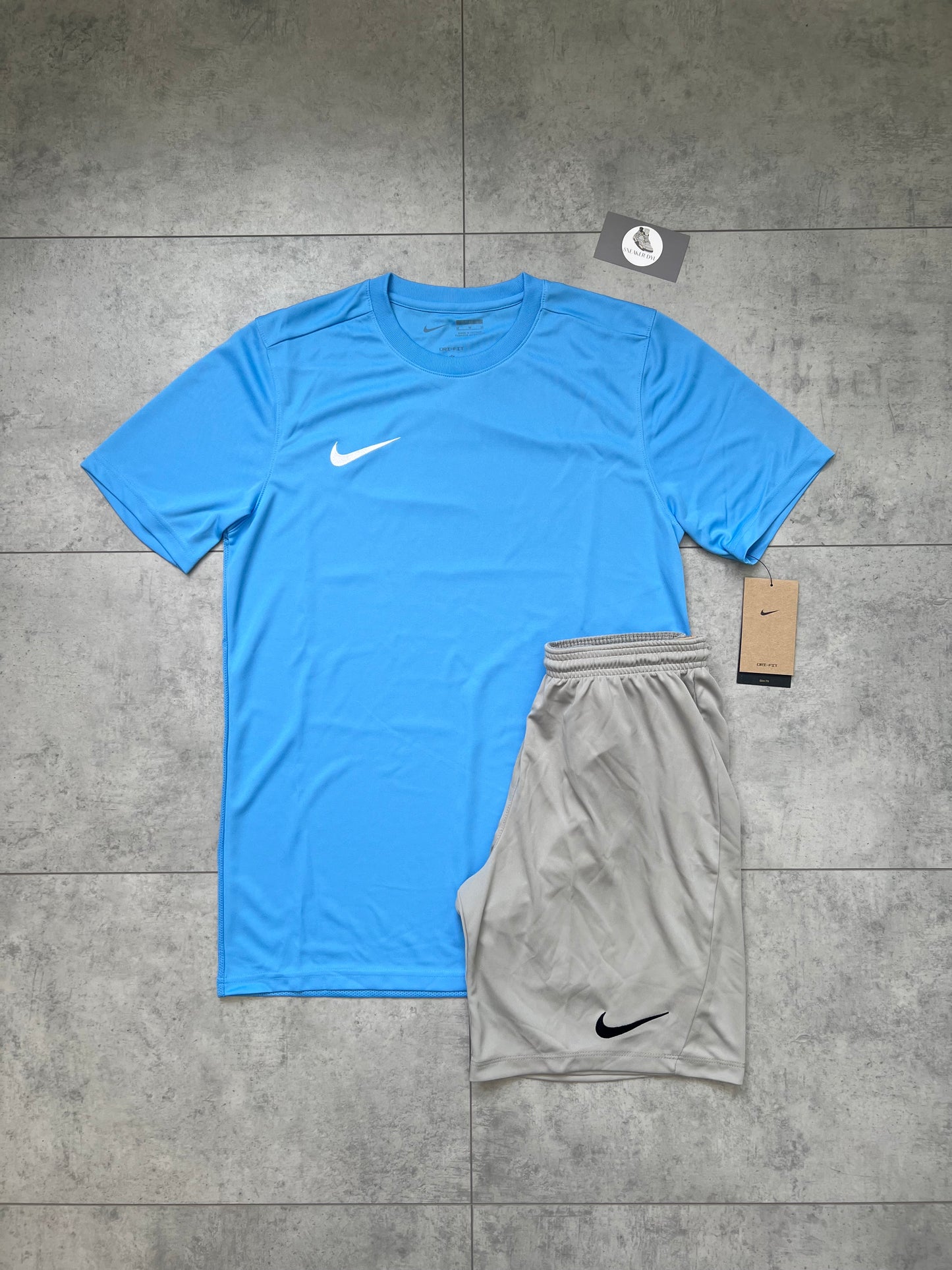 Flat-lay of Nike Dri Fit outfit set featuring a University Blue slim-fit t-shirt with light grey shorts, both with tags attached, laid out on a grey tiled background.