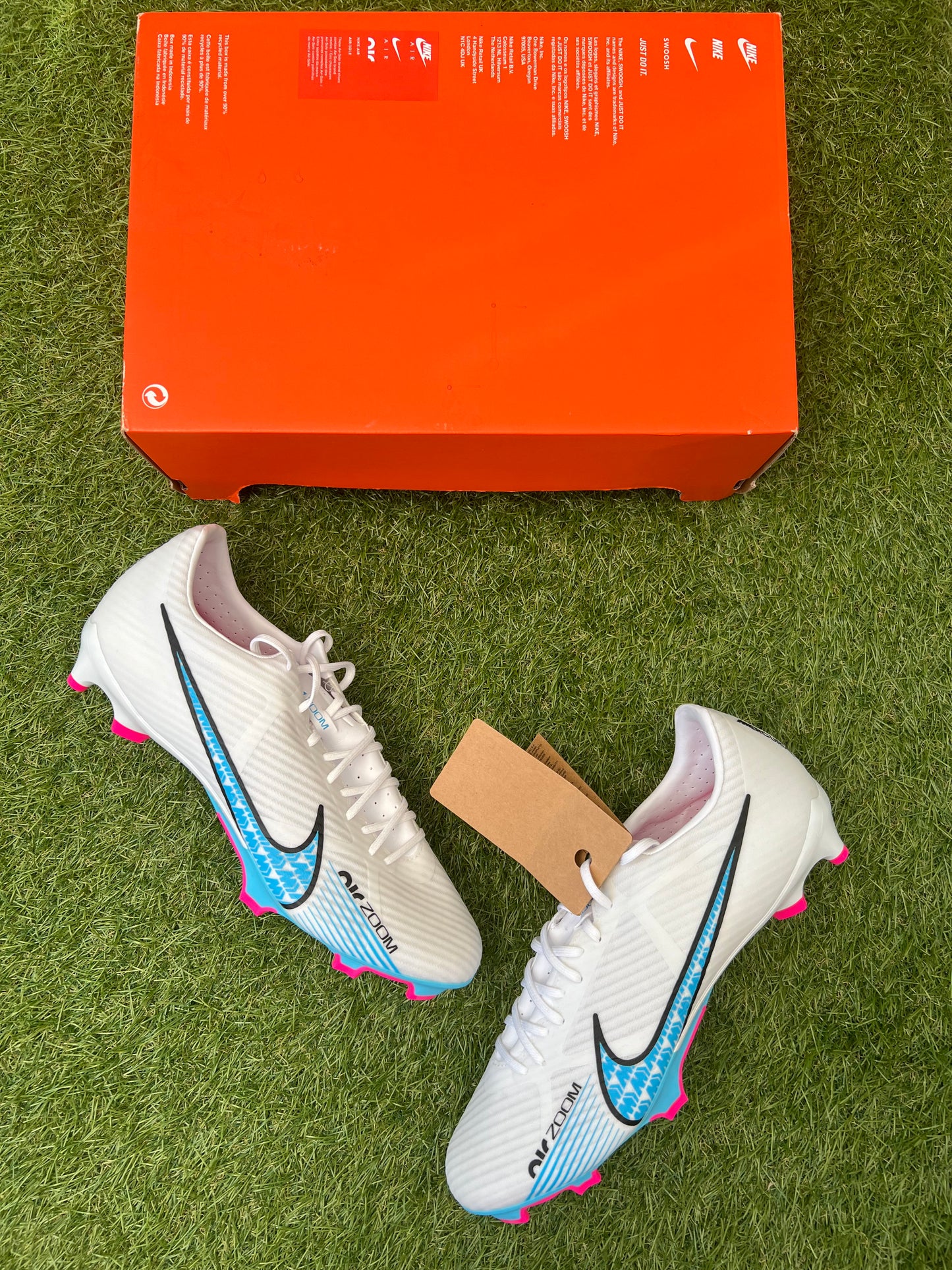 Nike Air Zoom Mercurial Vapor 15 Academy FG/MG football boots in white, Baltic Blue, and Hot Punch – lightweight synthetic upper with bold pink Swoosh and blue soleplate. Displayed on grass background, available at Sneaker Dyl.
