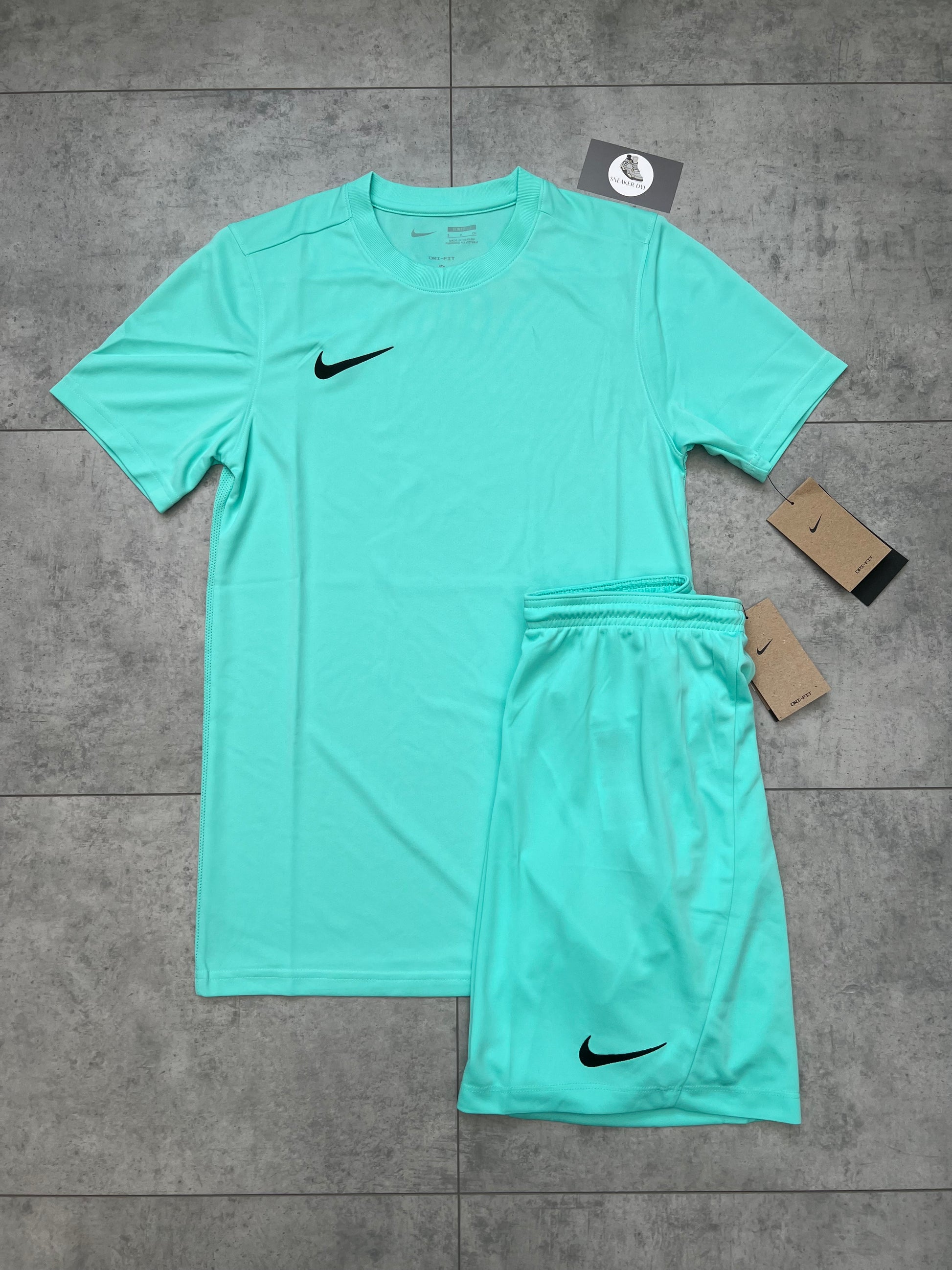 Flat-lay image of a brand new Nike Dri Fit Short & T-shirt Set in Hyper Turquoise. The set includes a slim-fit t-shirt and matching shorts with black Nike swoosh logos, displayed on a grey tiled background with product tags visible.